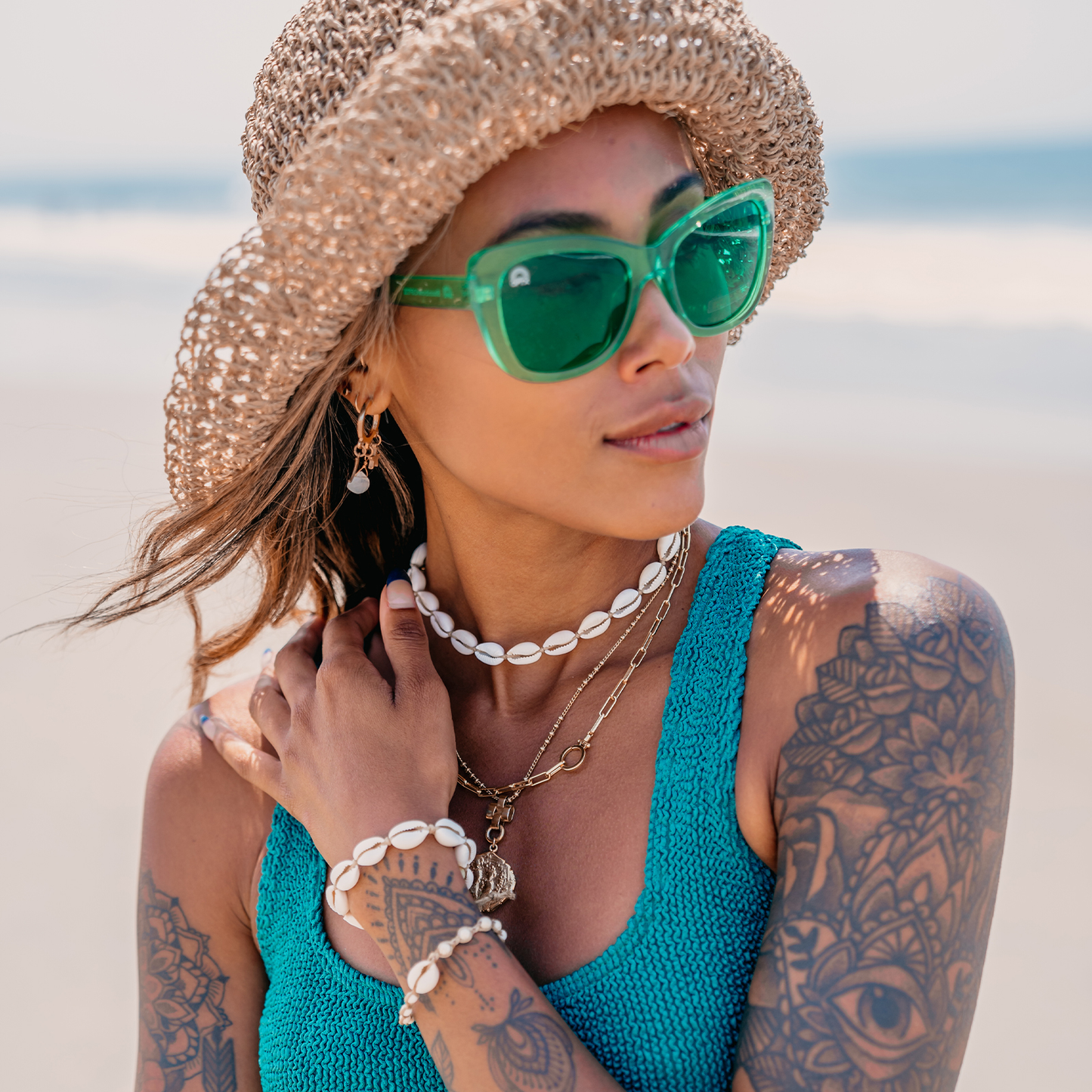 Woman wearing green sunglasses, a straw hat, and a blue top on a beach.