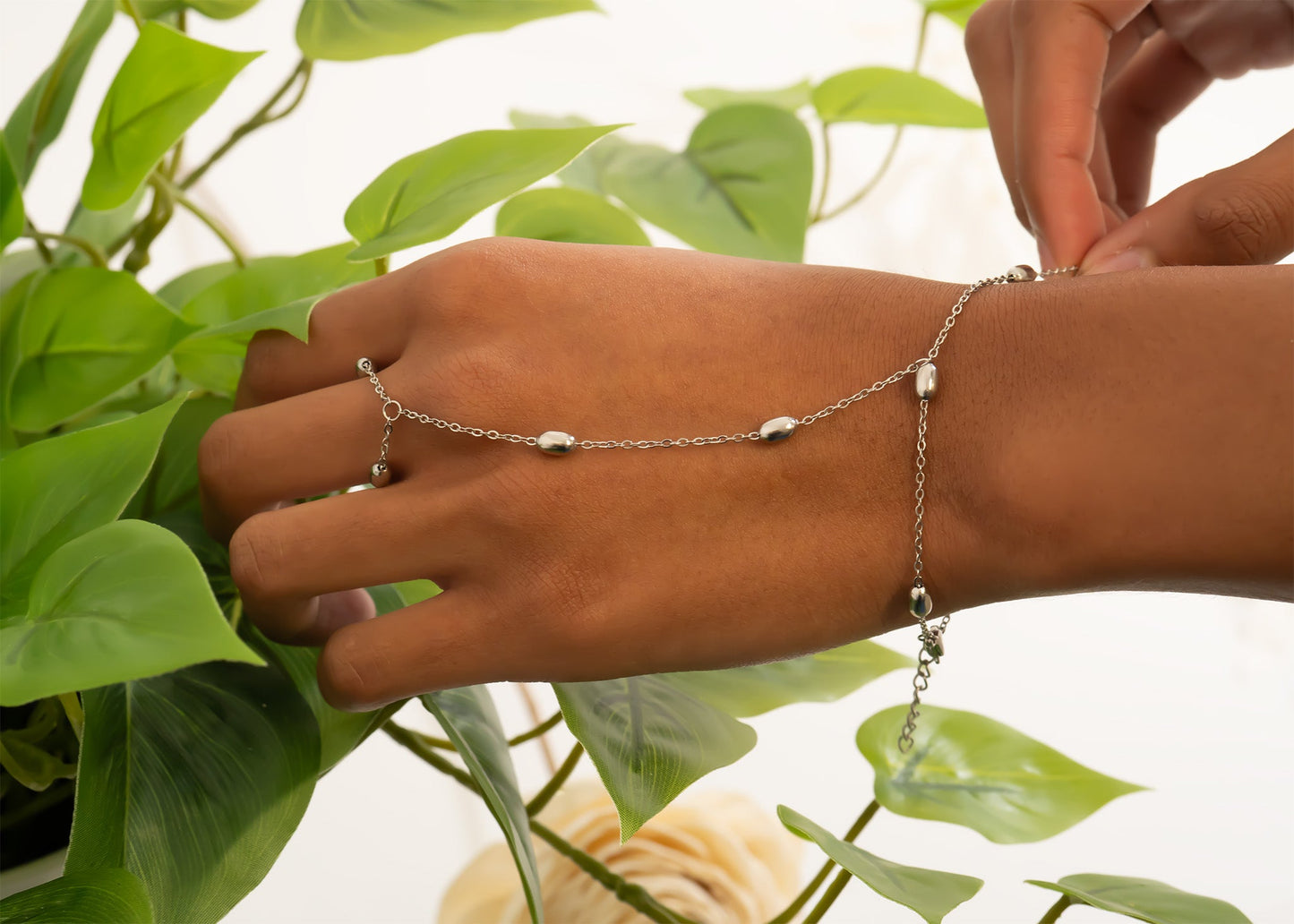Hand wearing a silver hand chain with green leaves in the background