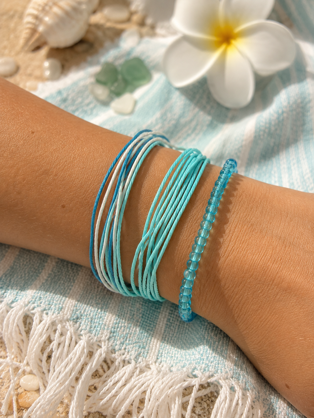 A close-up of a person's wrist wearing a stack of thin string bracelets in light blue, turquoise, and white, styled with a white frilly flower and seashells on a blue-and-white striped towel.