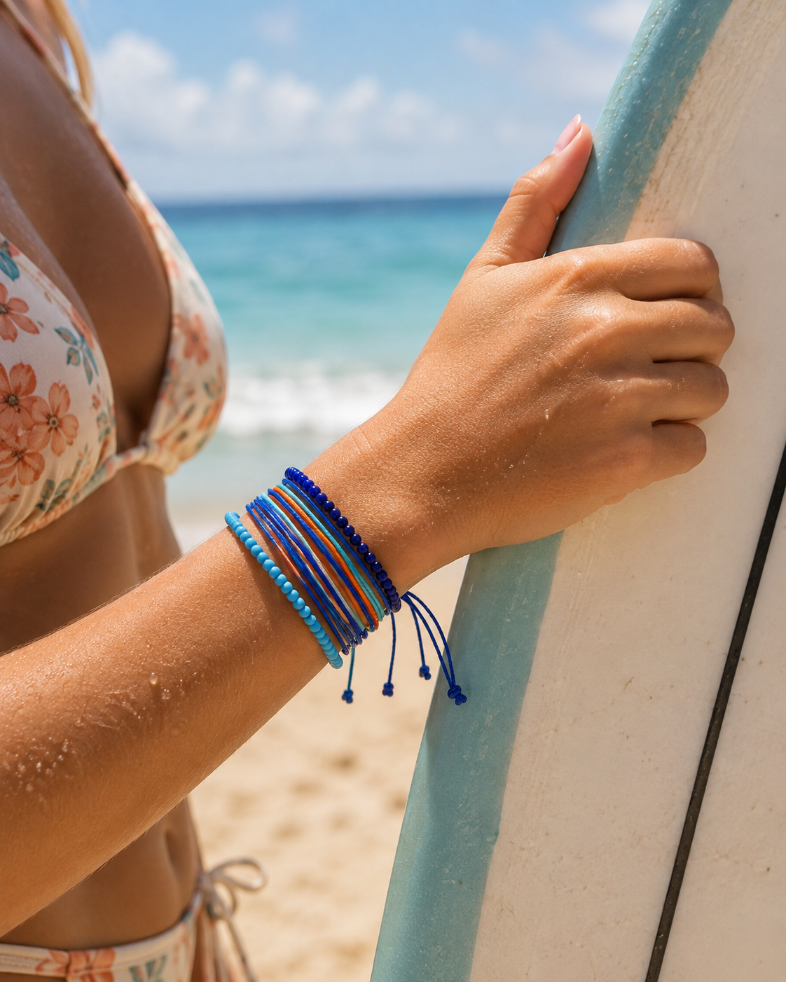 A close-up of a person's wrist wearing a stack of colorful braided and beaded bracelets, holding a surfboard on a sunny beach with the ocean in the background.