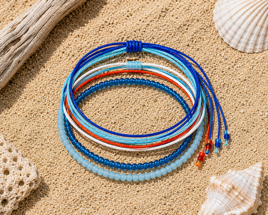 A stack of five colorful beaded and cord bracelets arranged in concentric circles on a sandy surface with seashells and driftwood props.