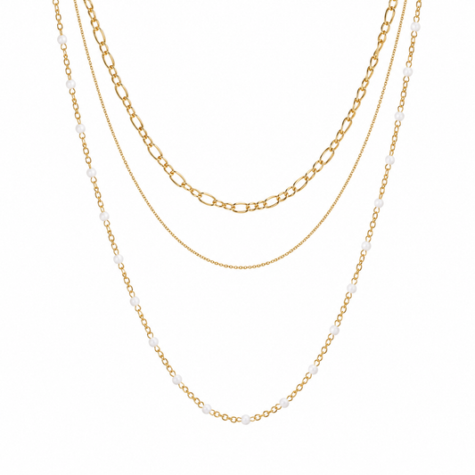 Three layered gold-tone necklaces displayed against a solid white background. The necklaces include a beaded chain with small white beads, a figaro chain, and a delicate cable chain.