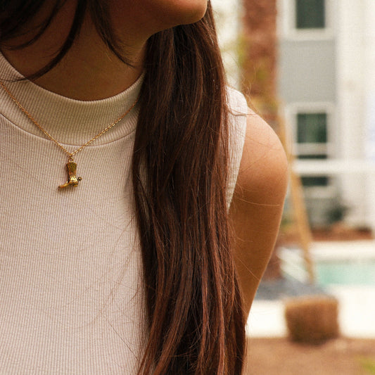 Girl with long, brown hair wearing a beige tank top wearing a gold chain with a cowboy boot pendant