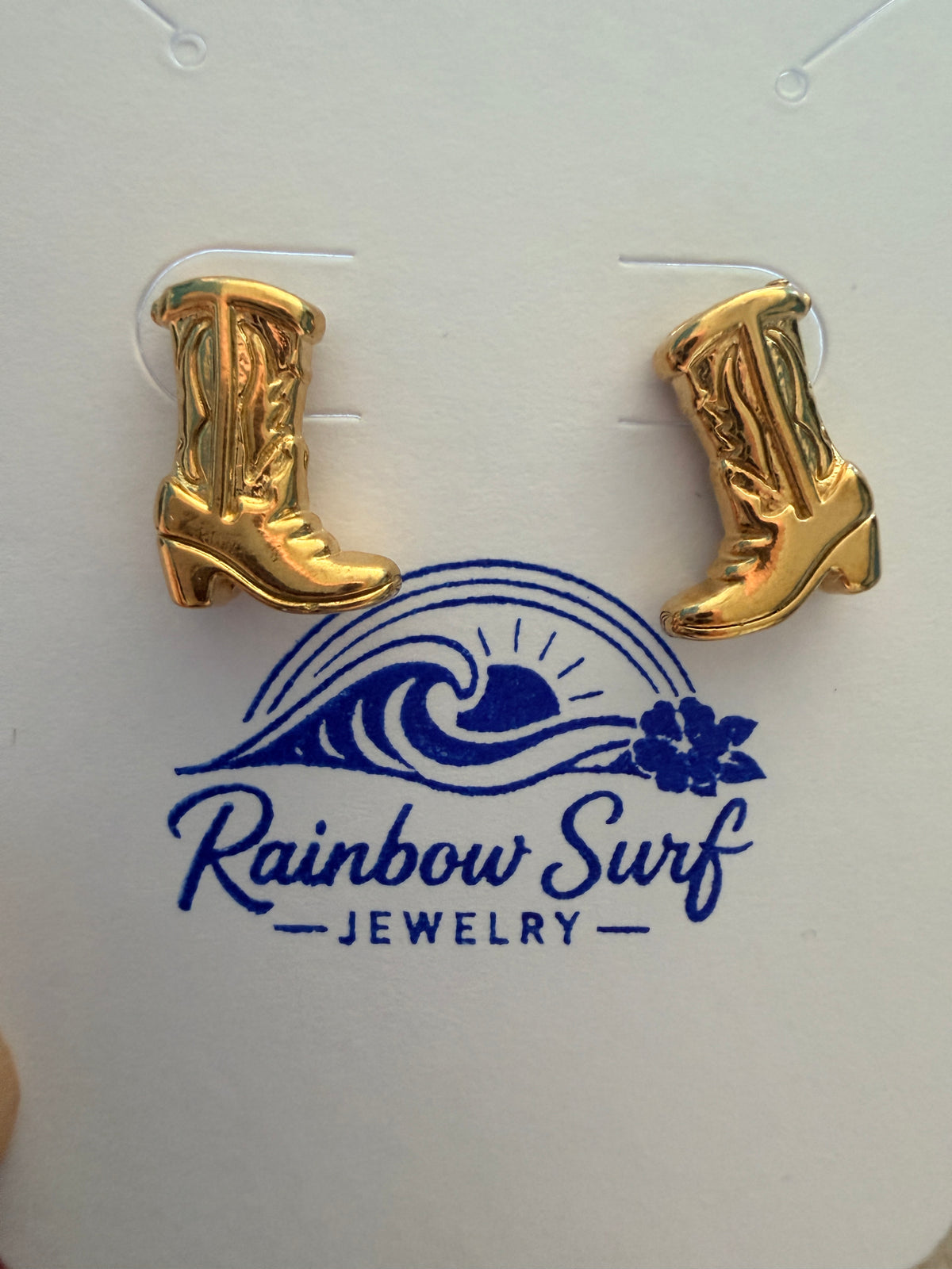 A pair of gold-colored cowboy boot stud earrings displayed on a white backing card with the blue 'Rainbow Surf Jewelry' logo.
