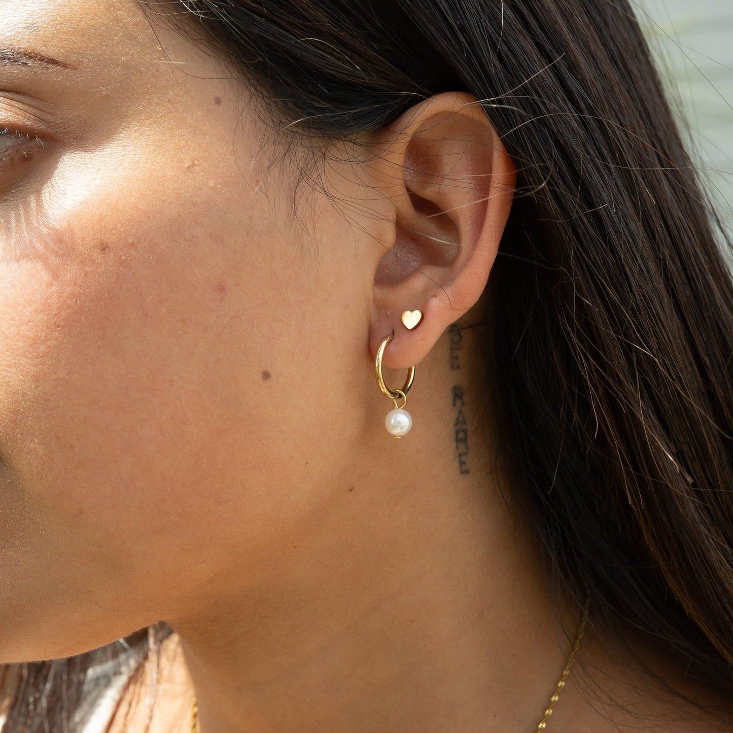 Close-up of a person wearing a gold hoop earring with a pearl and a second earring that is a heart shaped stud