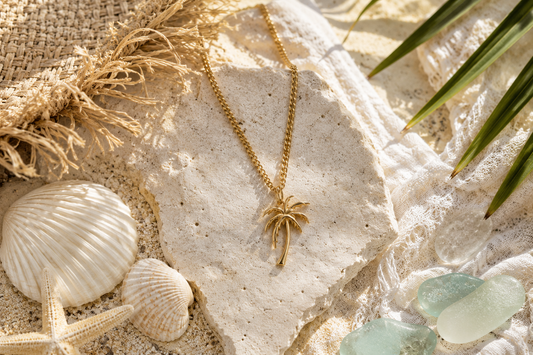 Gold chain with palm tree pendant lying next to a straw hat, sea glass, palm leaves, a starfish and seashells.