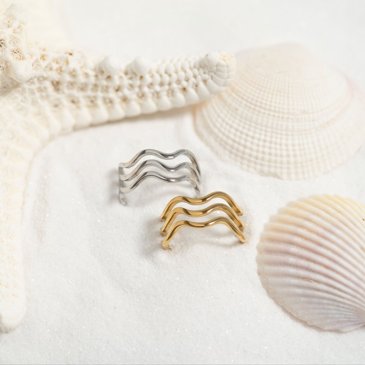 Silver and gold wave-shaped rings on a sandy surface with shells.