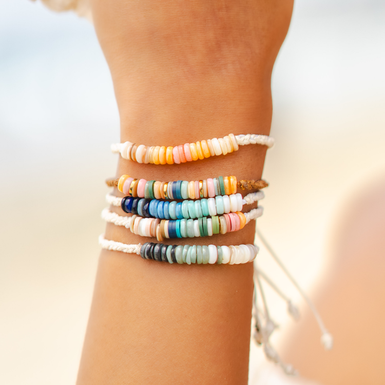 Close up of a hand with three bracelets, one pearl and one gold with a surfboard, palm trees and the beach in the background