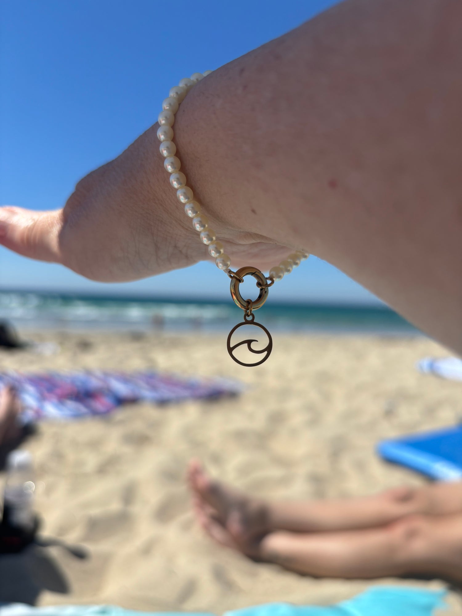 Person's arm stretched out in front of them wearing a pearl bracelet with a gold clasp and a gold ocean wave charm hanging from it with a beach background