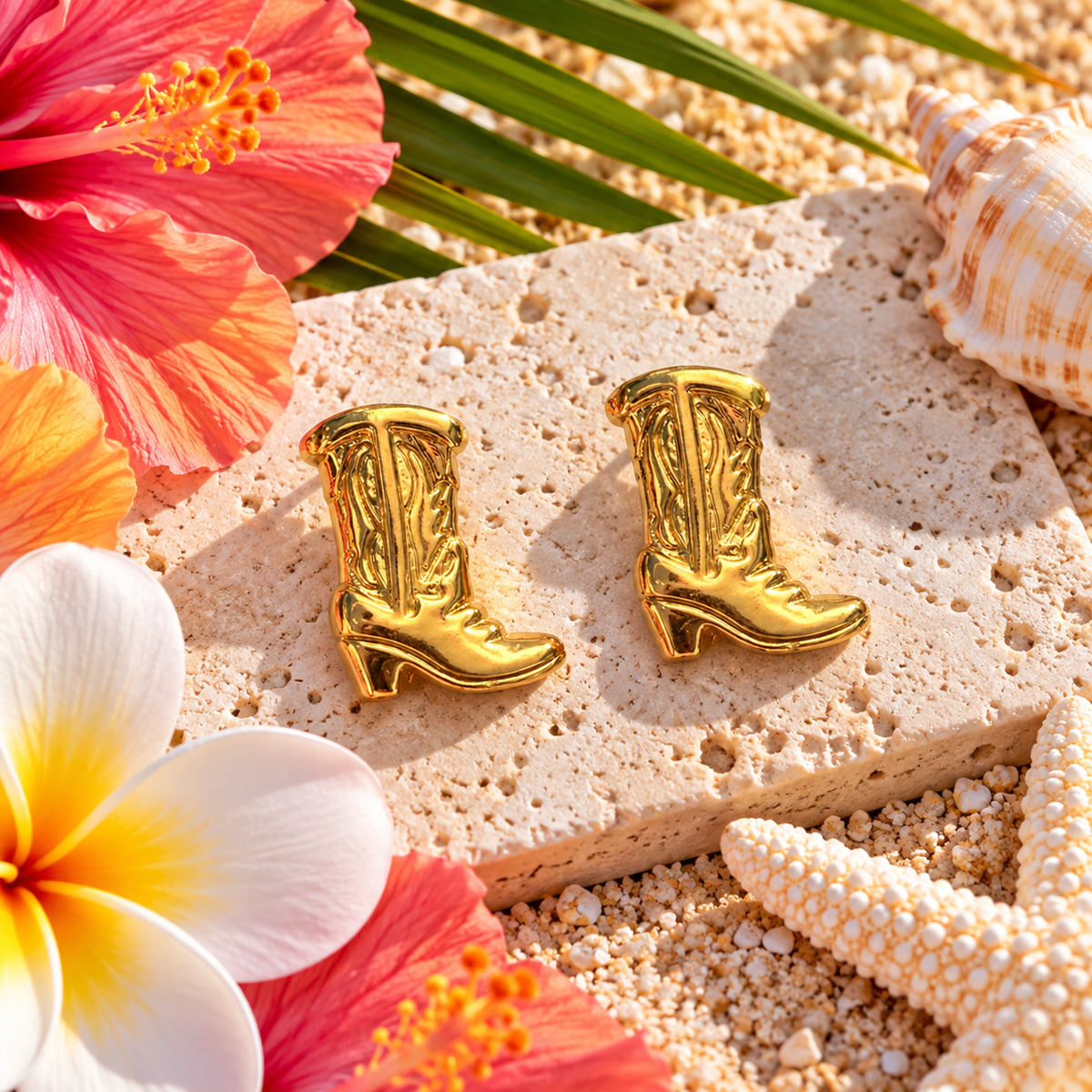 A pair of shiny gold cowboy boot stud earrings displayed on a textured stone slice over sand, surrounded by tropical flowers, a seashell, and a starfish.