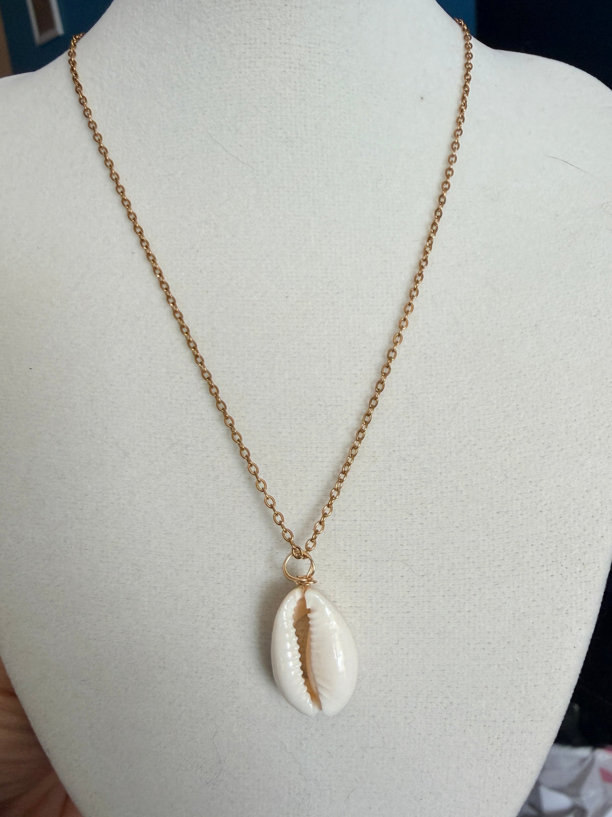 A gold chain necklace with a cowrie shell pendant displayed on a white mannequin bust. A hand is visible holding the left side of the mannequin stand.