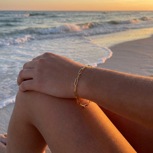 Hand wearing a gold paperclip chain bracelet on a beach with sunset