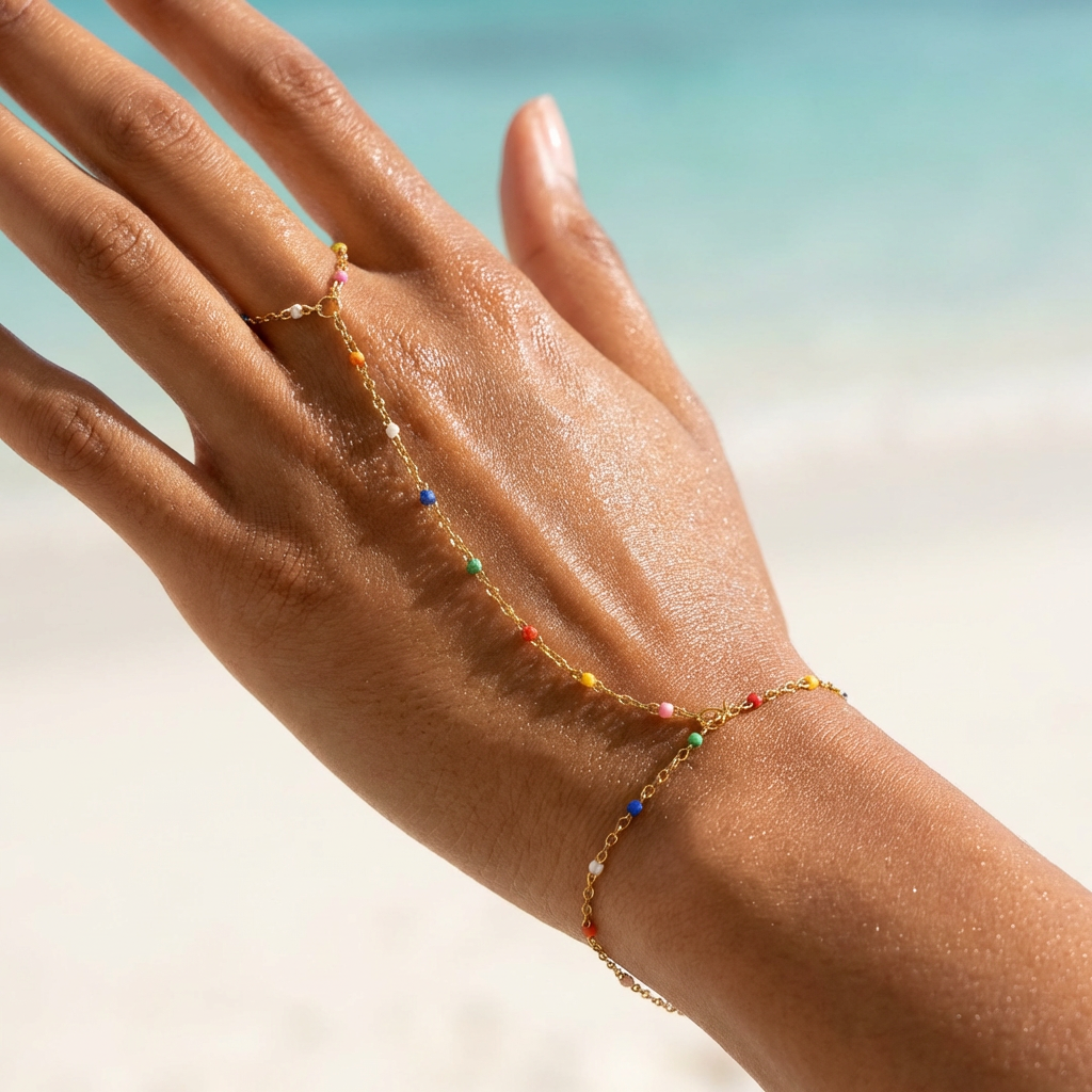 Hand wearing a gold hand chain with multi colored beads against a blurred beach background