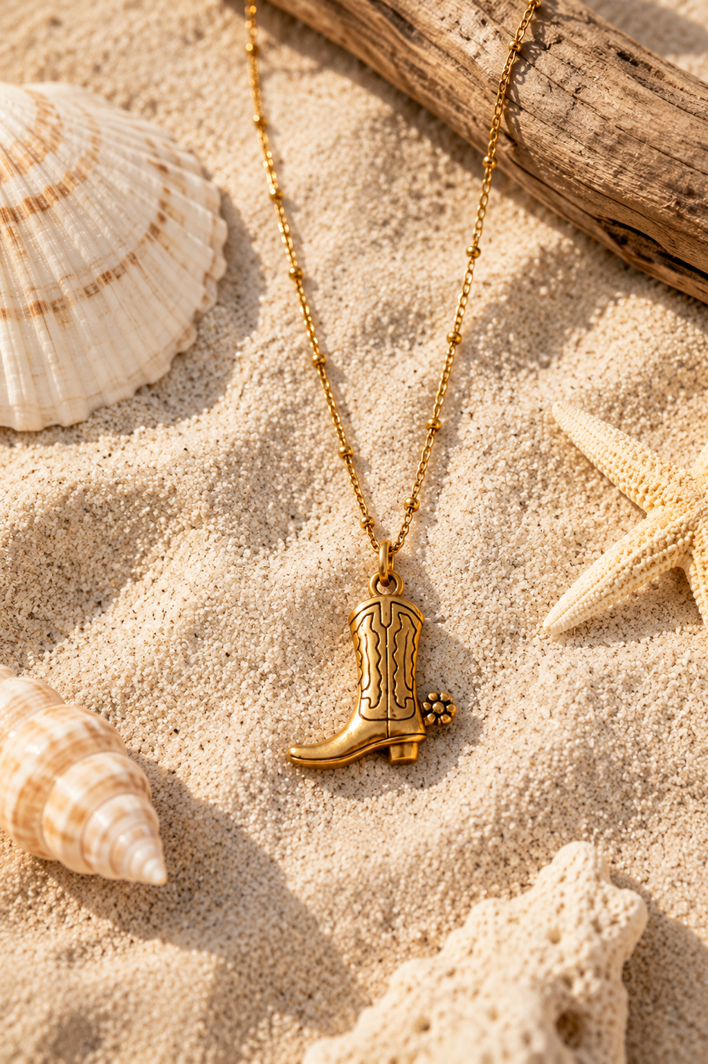 Gold satellite chain with cowboy boot pendant lying in sand surrounded by seashells.
