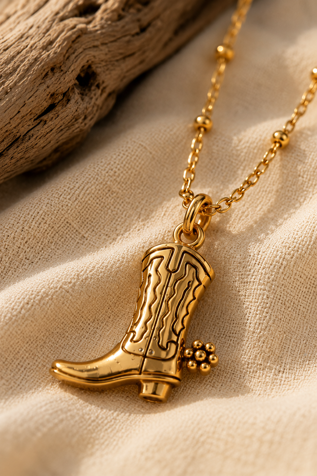 Gold satellite chain with cowboy boot pendant lying on cream-colored cloth.