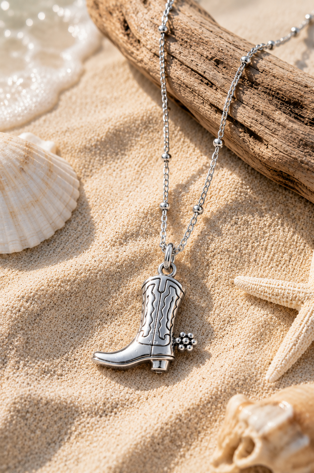 Silver satellite chain with cowboy boot pendant lying in the sand over a log surrounded by seashells.