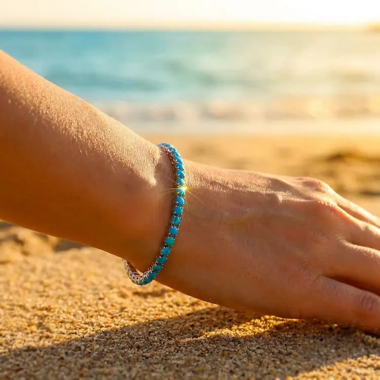 Hand wearing a blue beaded bracelet on a sandy beach with ocean in the background