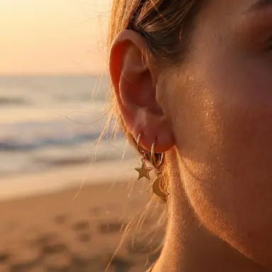 Close-up of an ear with gold crescent moon and star shaped earrings against a beach sunset background