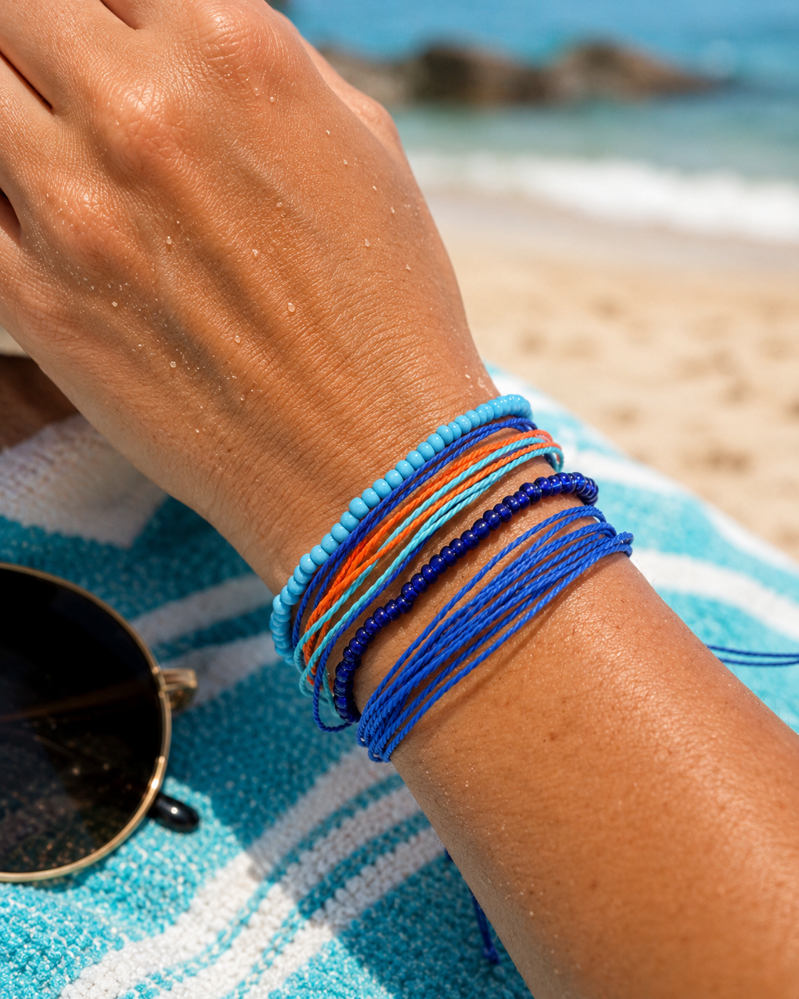 A close-up of a person's wrist wearing a stack of colorful beaded and cord bracelets on a beach, with a blue and white striped towel and sunglasses in the foreground.