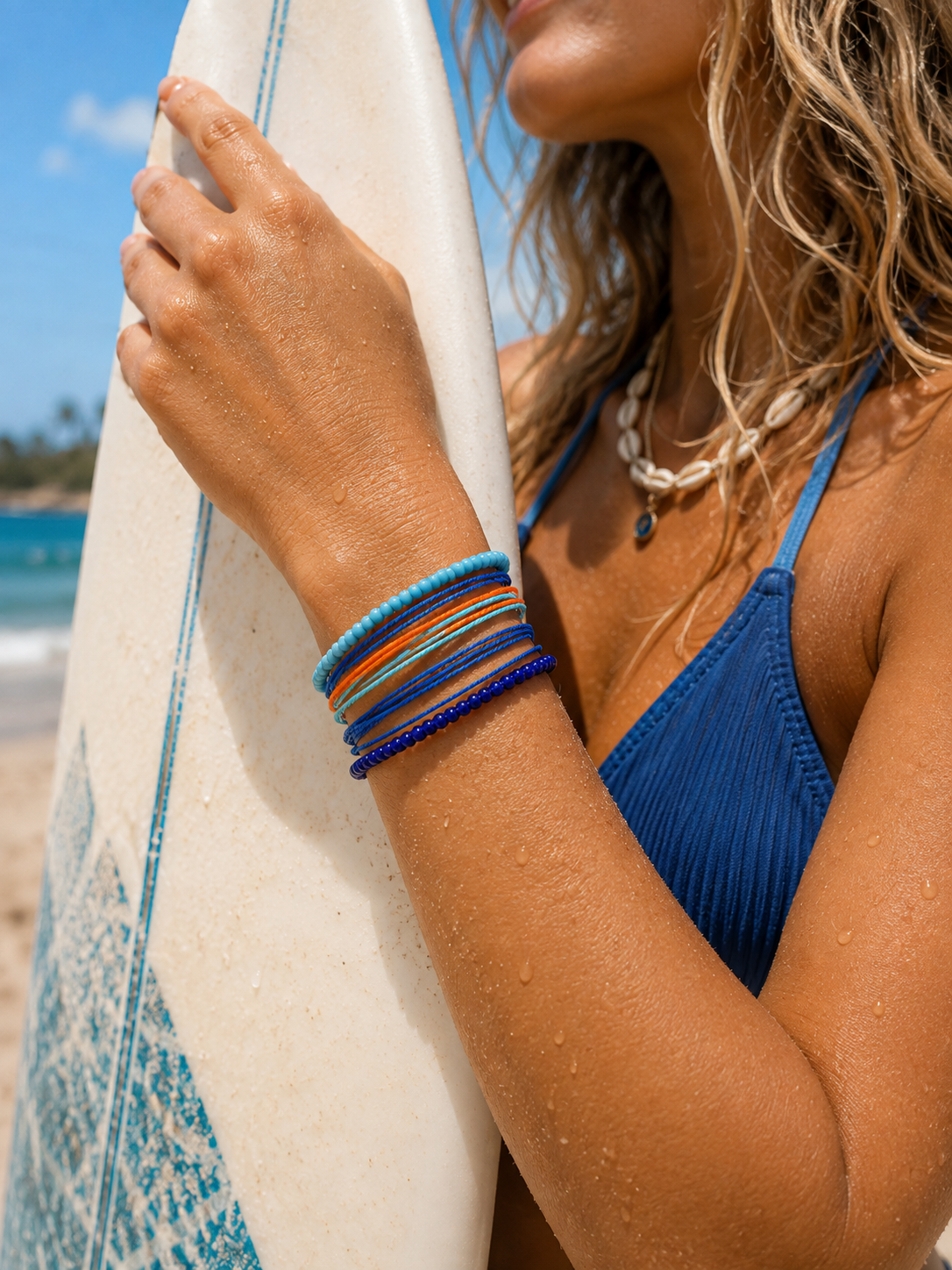 A close-up lifestyle shot of a woman on a beach wearing a stack of colorful beaded bracelets, holding a white surfboard with blue patterns. She is wearing a blue bikini top and a shell necklace.