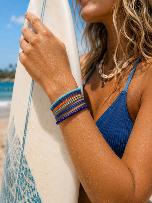 A close-up lifestyle shot of a woman on a beach wearing a stack of colorful beaded bracelets, holding a white surfboard with blue patterns. She is wearing a blue bikini top and a shell necklace.