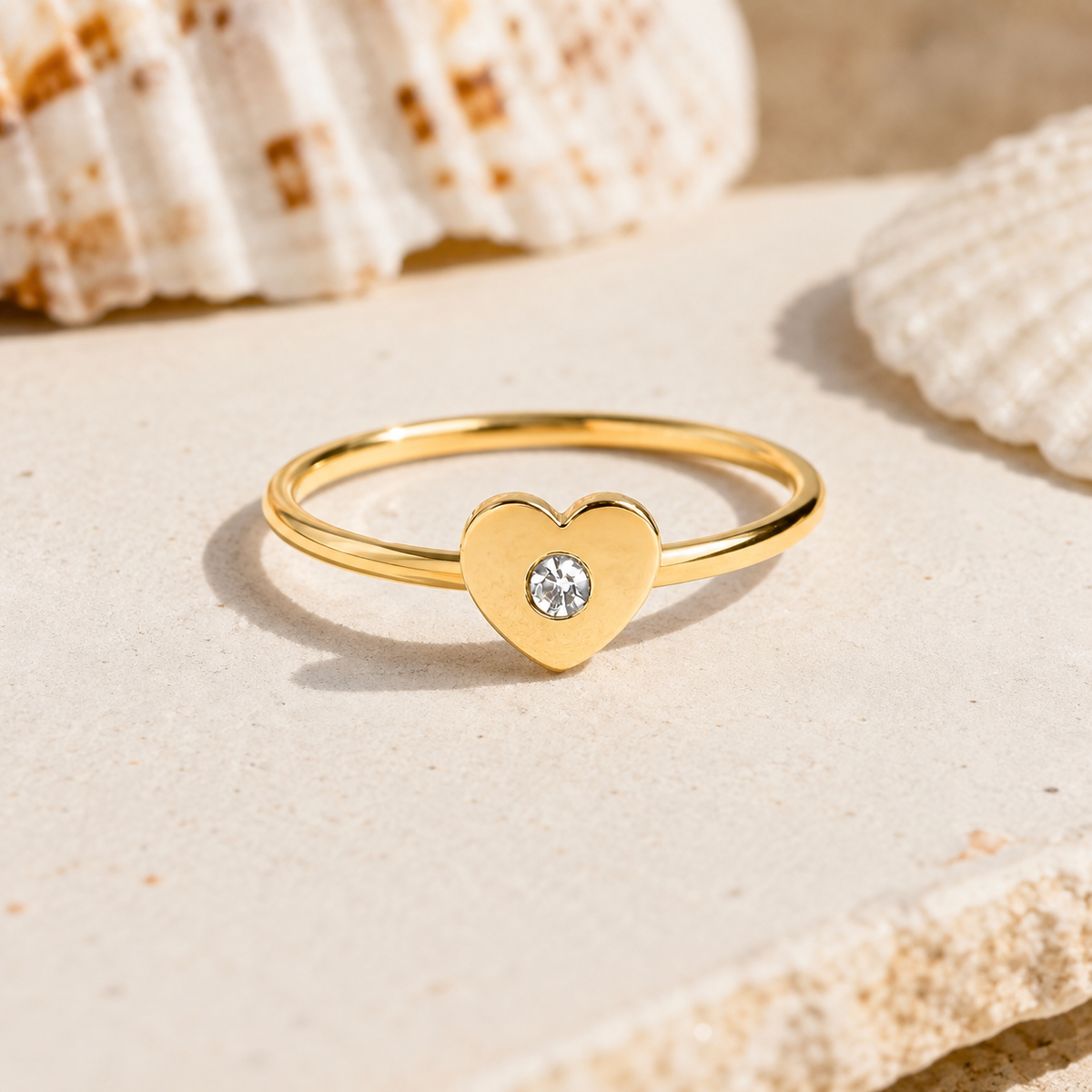 A gold ring with a heart-shaped bezel holding a clear round gemstone, displayed on a light stone surface with seashells in the background.