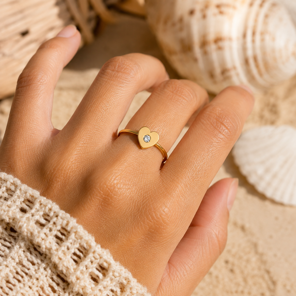 A gold heart ring with a small clear gemstone is worn on a person's finger, set against a sandy beach background with seashells and a woven basket.