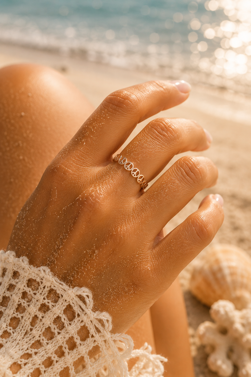 A close-up of a person's hand resting on a sandy beach, wearing a delicate gold ring with a repeating wave pattern. The background features the ocean water and a soft-focus seashell.