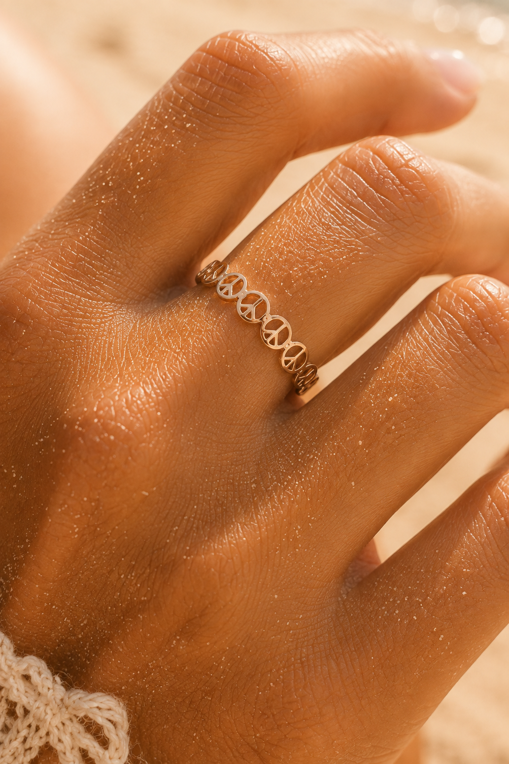 A close-up of a person's hand wearing a delicate gold ring with a continuous interlocking infinity symbol design, set against a warm, sandy beige background.