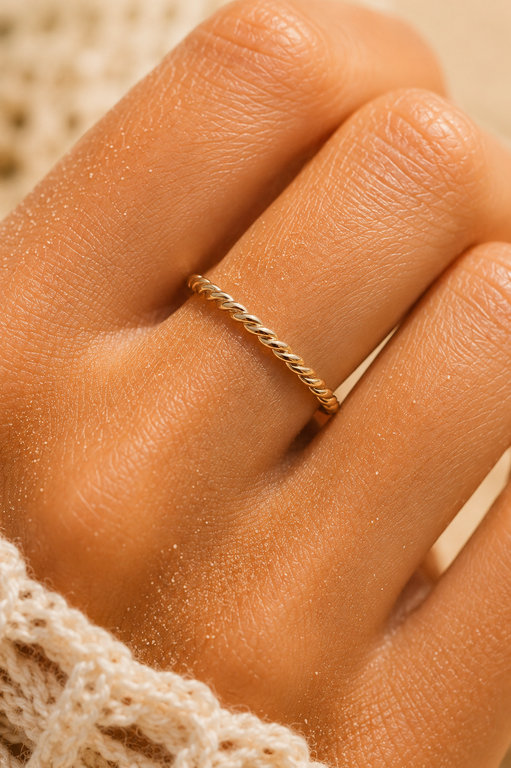 A close-up of a hand wearing a thin twisted gold band ring, with a cream-colored knit sleeve visible at the wrist.