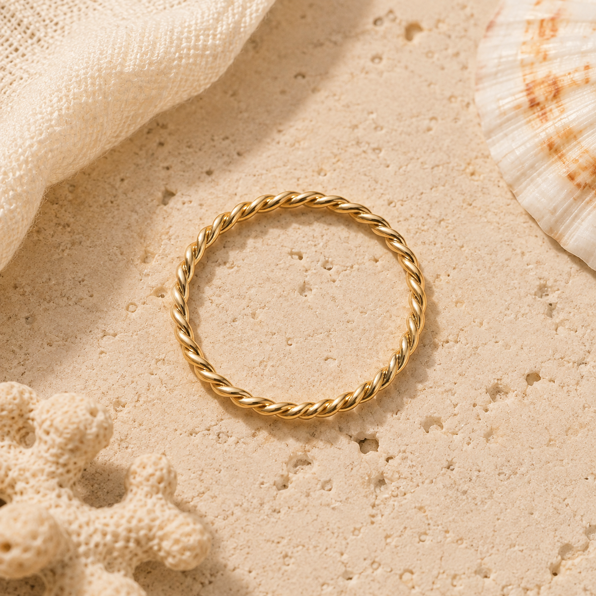 A thin gold twisted rope ring lies on a textured beige stone surface with decorative seashell and coral props in the corners.