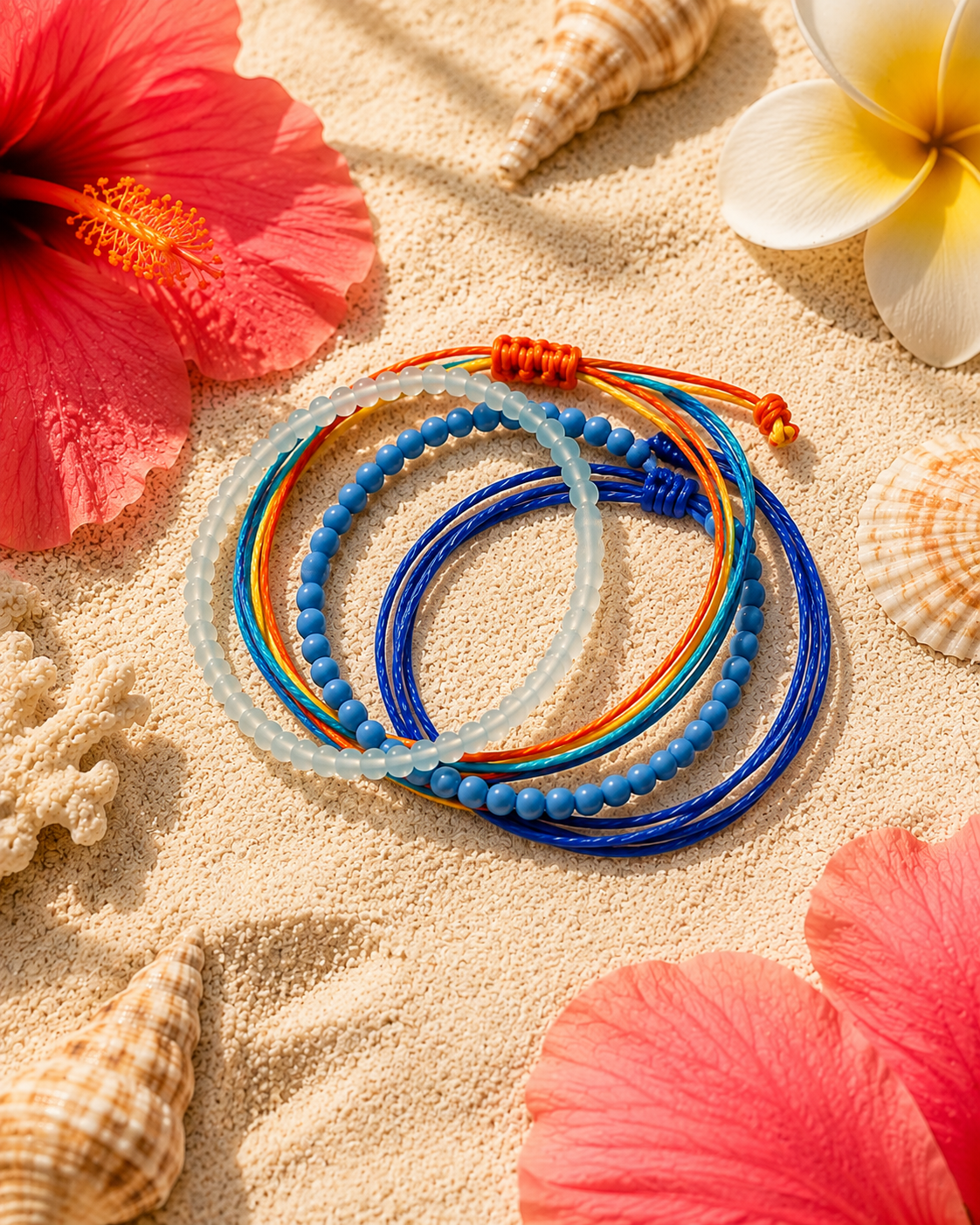 Four colorful beaded and cord bracelets arranged on sand with seashells and hibiscus flowers. The bracelets include translucent pale blue, medium blue, royal blue, and orange with blue accents.
