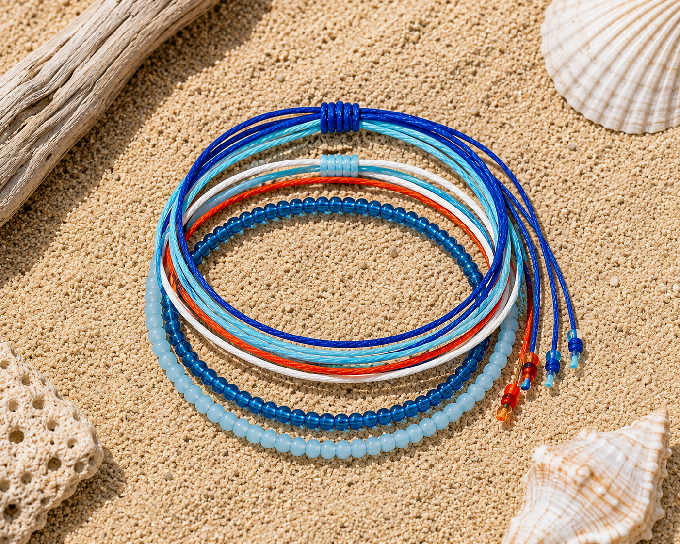 A stack of five colorful beaded and cord bracelets arranged in concentric circles on a sandy surface with seashells and driftwood props.