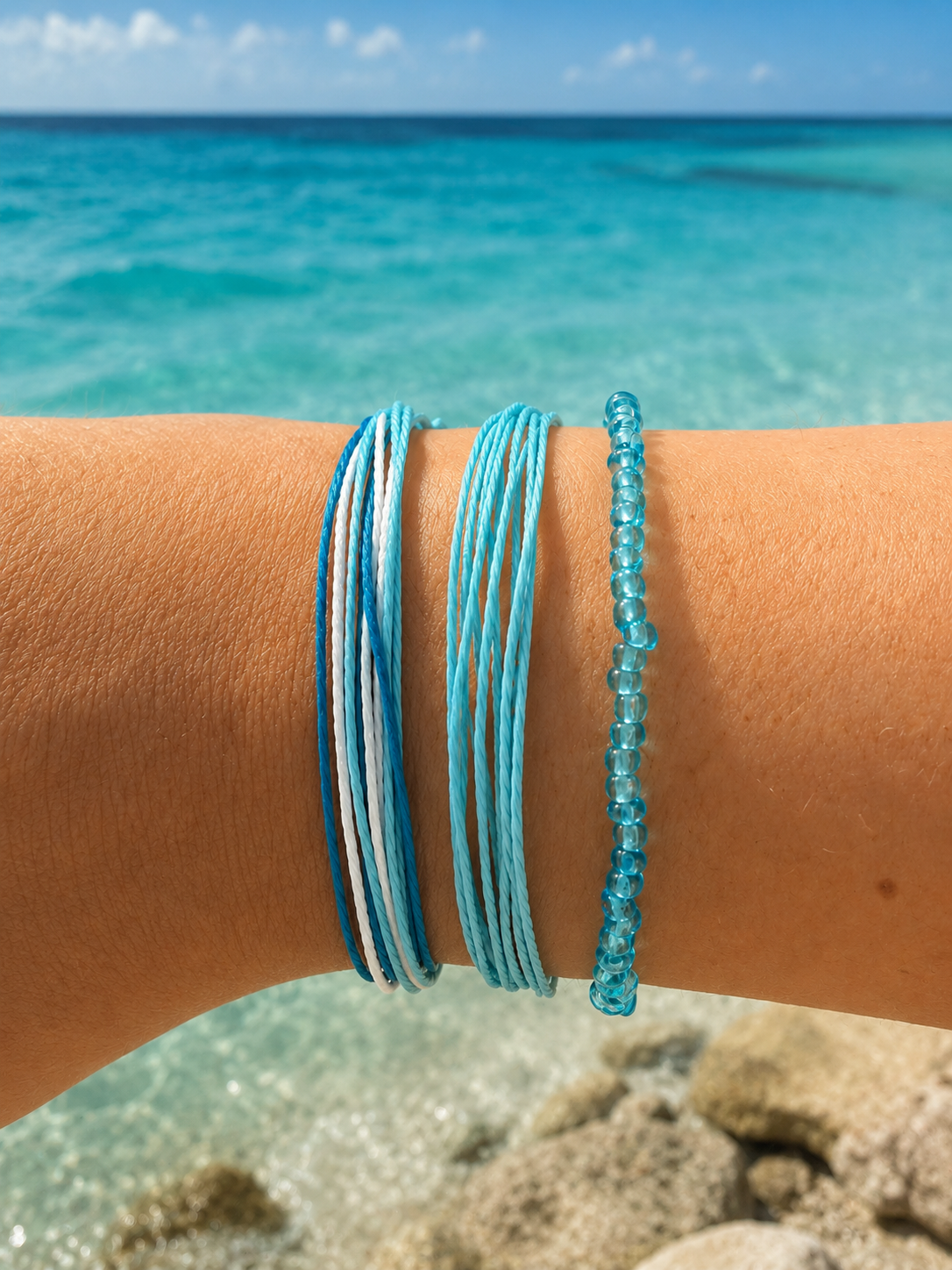 Three blue and white bracelets worn on a person's wrist, with a blurred tropical beach and turquoise ocean in the background.