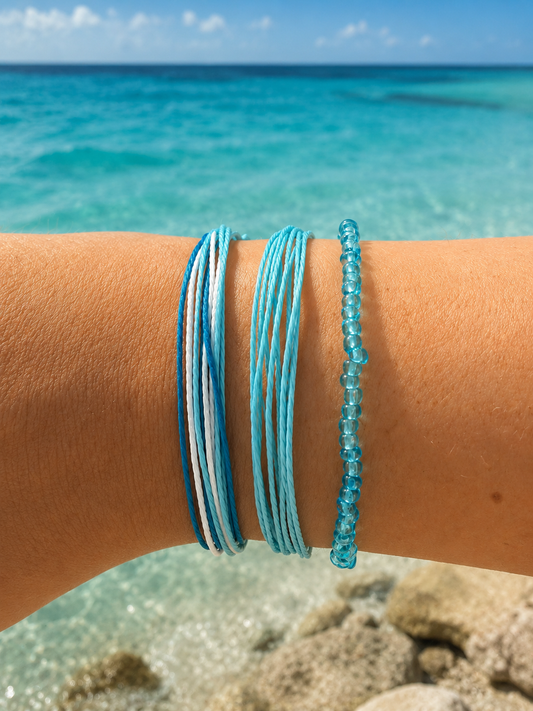 Three blue and white bracelets worn on a person's wrist, with a blurred tropical beach and turquoise ocean in the background.