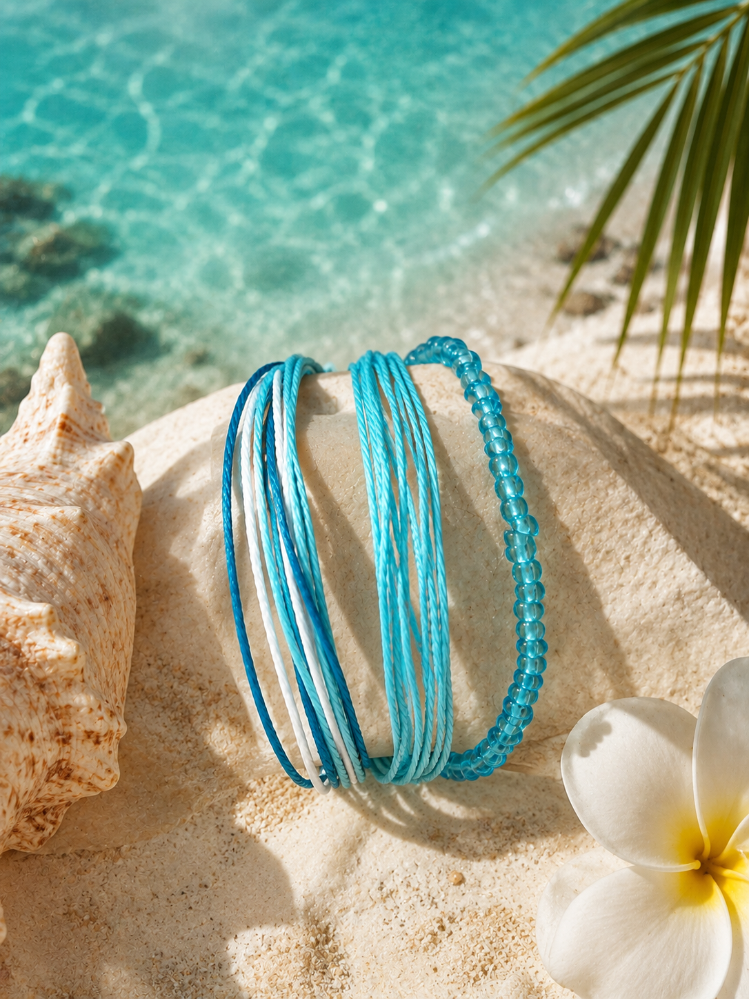 A set of turquoise and blue beaded and cord bracelets displayed on a sand-covered shell, styled with a seashell, white flower, and a tropical ocean background.