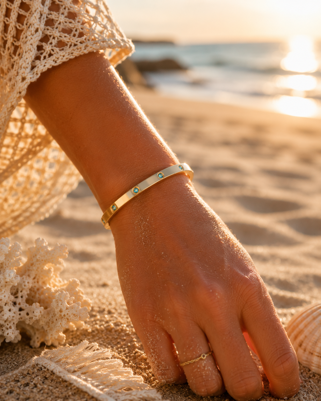 A gold hinged bracelet with small turquoise stone accents worn on a woman's wrist, shown in a close-up beach lifestyle shot with sand, a seashell, and a sunset background.