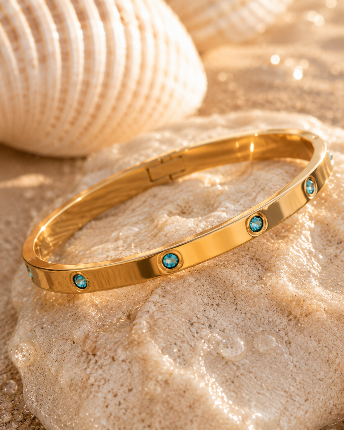 A polished gold hinged bracelet with small round turquoise-blue gemstone accents, resting on a textured beige stone with a seashell in the background.