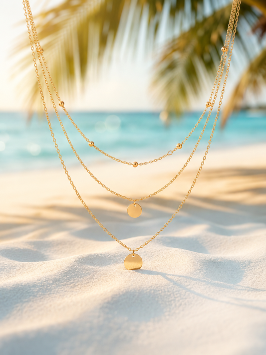 Three layered gold chain necklaces with small disc pendants, displayed against a tropical beach background with sand, ocean, and palm leaves.
