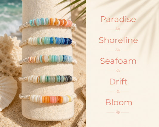 Five beaded bracelets in pastel and ocean-inspired colors displayed on a cream cylindrical stand, set on sand with a shell and tropical water background. Text on the right lists the bracelet names: Paradise, Shoreline, Seafoam, Drift, and Bloom.