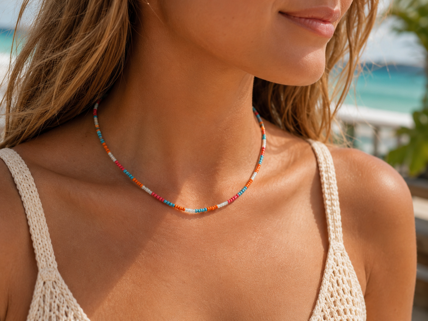A close-up lifestyle shot of a woman wearing a delicate beaded necklace with orange, blue, and white beads, paired with a cream crochet bikini top against a blurred beach background.