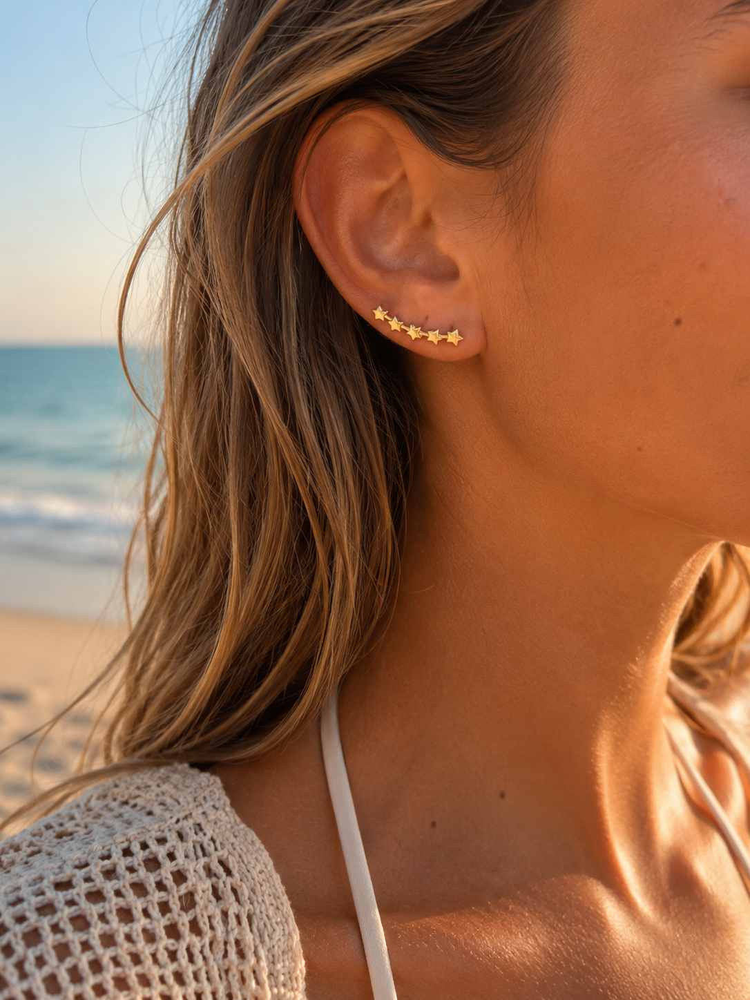 A close-up of a woman's ear at a beach wearing a small gold ear cuff with five star-like studs.