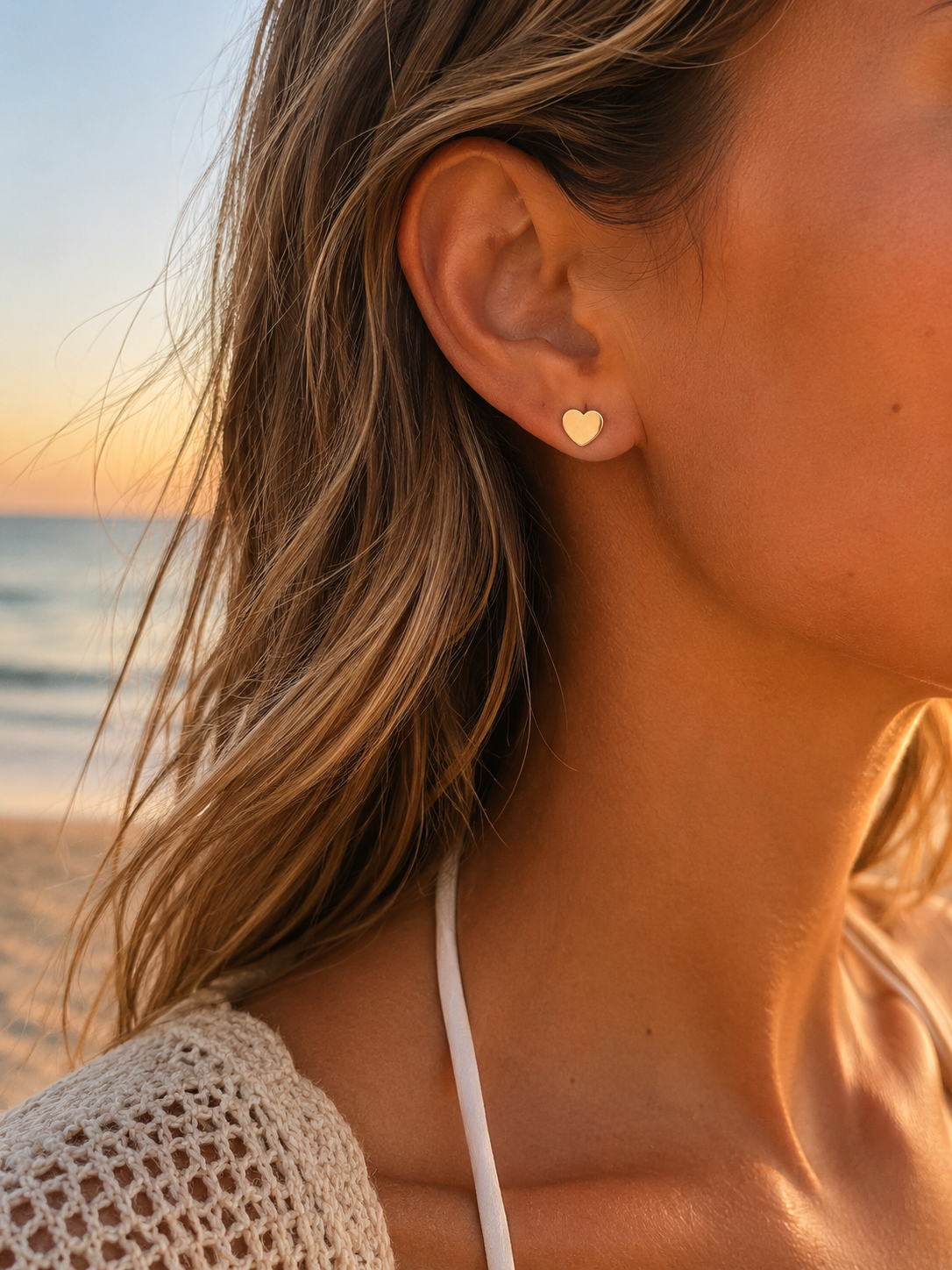 A close-up side profile of a woman on a beach wearing a small gold heart stud earring, with ocean waves and a sunset sky in the background.