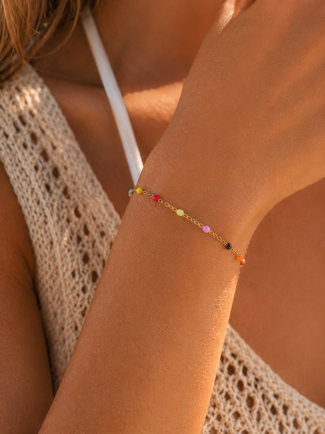 A delicate gold chain bracelet with small colorful beads worn on a woman's wrist, shown in a warm, sunlit close-up.