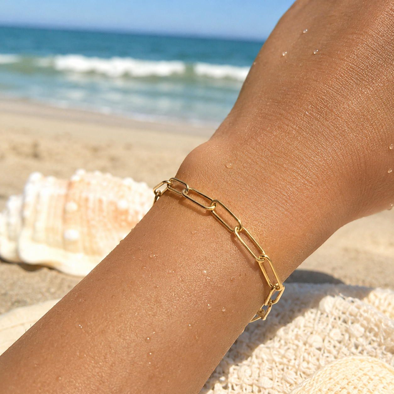 A close-up of a person's wrist on a beach wearing a delicate gold paperclip chain bracelet, with sand, a seashell, and the ocean in the background.