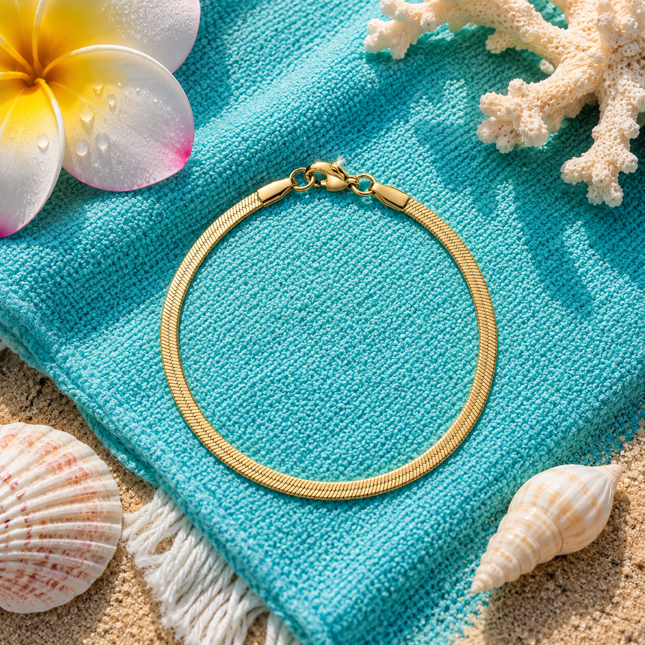 A gold-toned herringbone chain bracelet arranged in a circle on a turquoise beach towel, styled with seashells, coral, and a plumeria flower on sand.