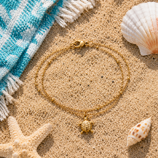 A gold double-strand chain anklet with a turtle charm, styled on a sandy beach flat-lay with a blue and white towel, starfish, and seashells.