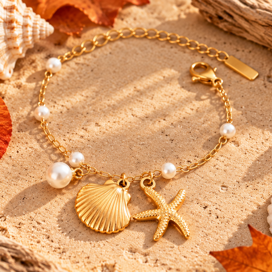 A gold-tone chain bracelet with a lobster clasp, decorated with white pearl beads and two charms: a textured seashell and a starfish, displayed on a sandy beach background with seashells and autumn leaves.