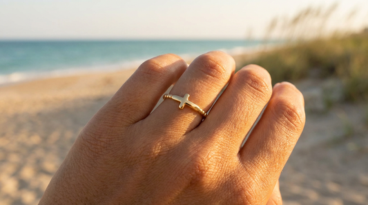 Hand wearing a gold cross ring on a beach with ocean and sand in the background
