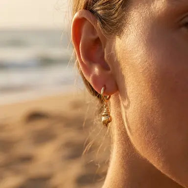 Close-up of a person's ear with a gold conch shell earring on a beach.